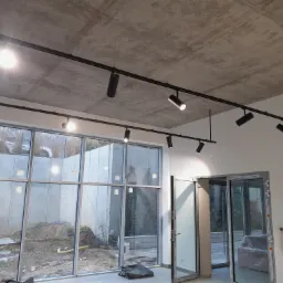 Nowoczesne oświetlenie szynowe z reflektorami LED w surowym wnętrzu z betonowym sufitem i dużymi oknami wychodzącymi na teren budowy.