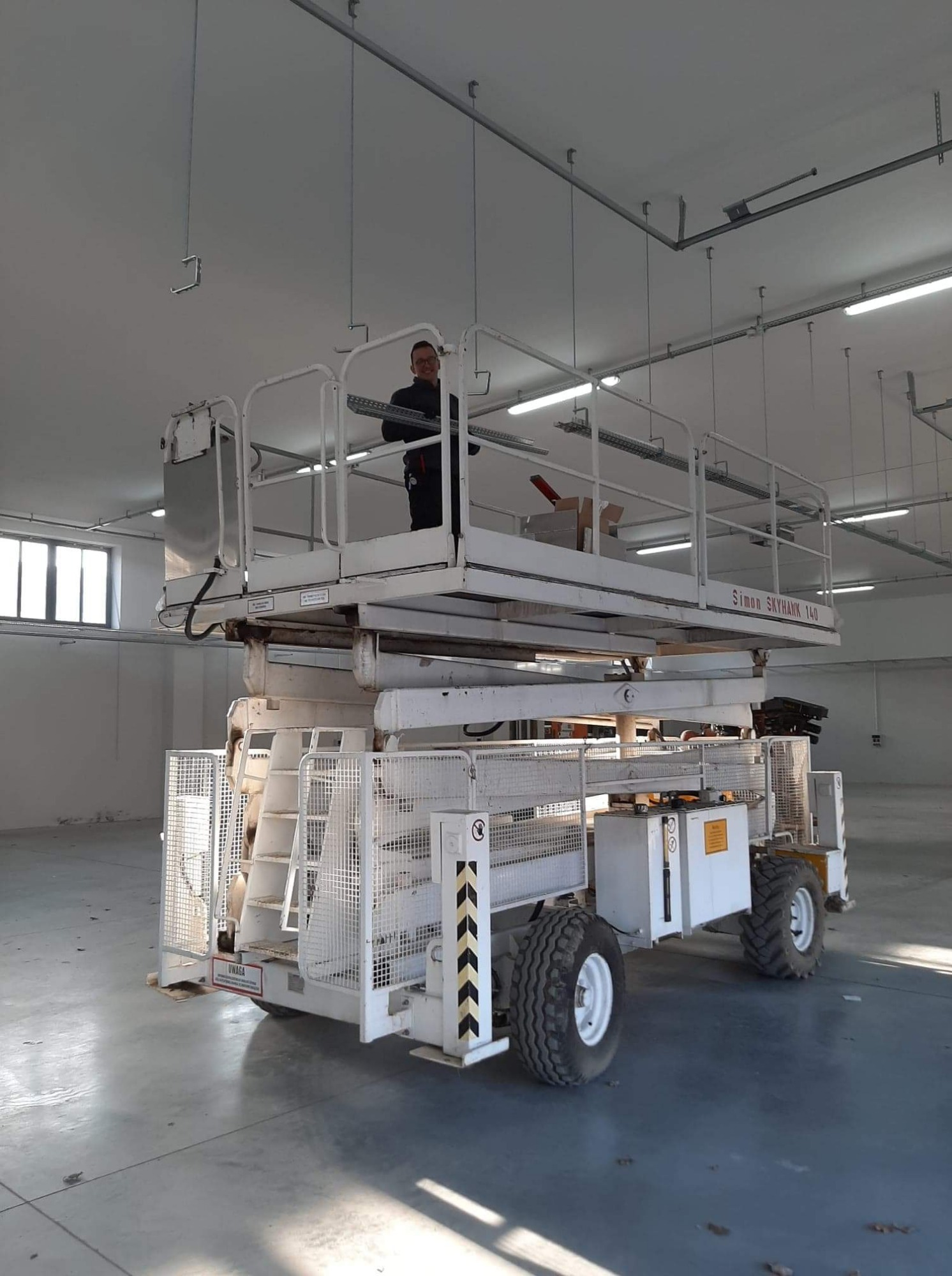 Pracownik na podnośniku nożycowym Simon Skyhawk 140, montujący elementy instalacji pod sufitem w hali.