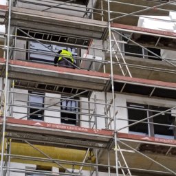 RESBUD A.J. SPÓŁKA Z OGRANICZONĄ ODPOWIEDZIALNOŚCIĄ - Budynek w trakcie prac elewacyjnych, widoczne rusztowania i pracownik w żółtej kamizelce. Częściowo ocieplona ściana z widocznym styropianem. Ujęcie z dołu.