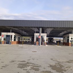 RESBUD A.J. SPÓŁKA Z OGRANICZONĄ ODPOWIEDZIALNOŚCIĄ - Nowoczesny punkt kontroli granicznej z szarym zadaszeniem i kostką brukową. Widoczne budki kontrolne z ceglanymi elementami i znakami zakazu wjazdu. Jasne niebo w tle.