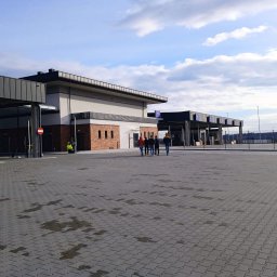 RESBUD A.J. SPÓŁKA Z OGRANICZONĄ ODPOWIEDZIALNOŚCIĄ - Rozległy plac wyłożony szarą kostką brukową, z budynkami w tle i grupą osób przechodzących przez przejście dla pieszych. Widoczne słupy oświetleniowe i błękitne niebo.