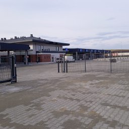 RESBUD A.J. SPÓŁKA Z OGRANICZONĄ ODPOWIEDZIALNOŚCIĄ - Rozległy plac wyłożony szarą kostką brukową, z budynkiem w tle. Widoczne bramy wjazdowe i szlabany. Całość w stonowanej kolorystyce, przy pochmurnym niebie.