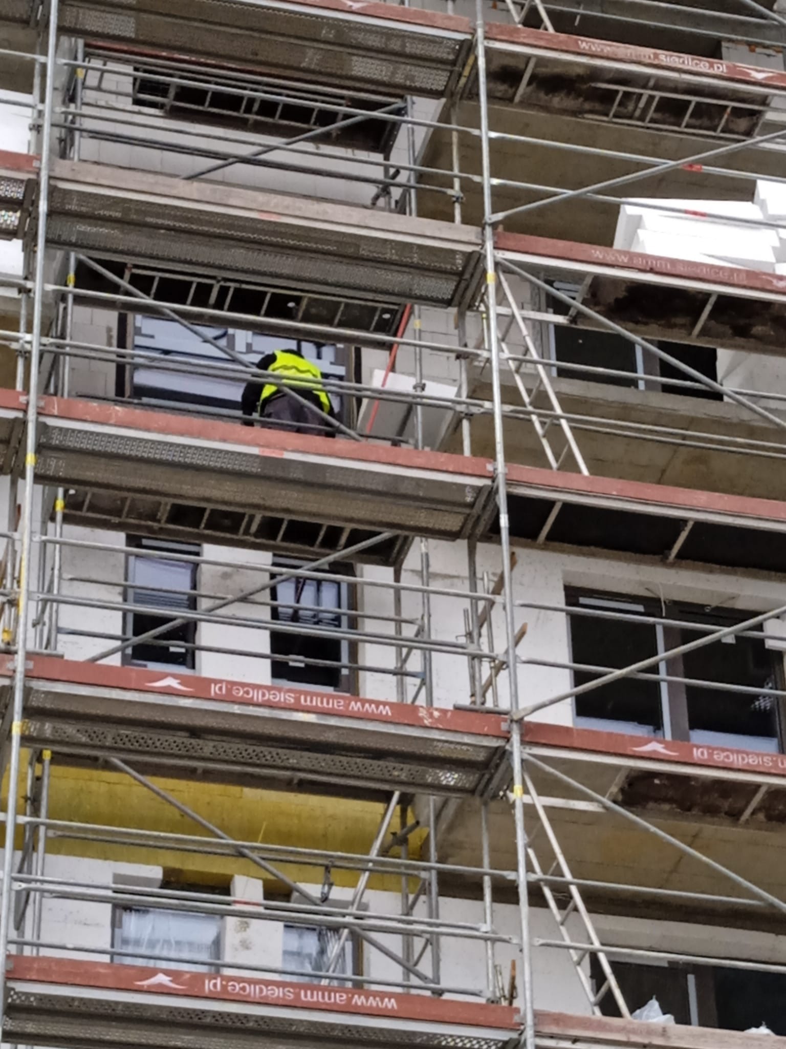 Budynek w trakcie prac elewacyjnych, widoczne rusztowania i pracownik w żółtej kamizelce. Częściowo ocieplona ściana z widocznym styropianem. Ujęcie z dołu.