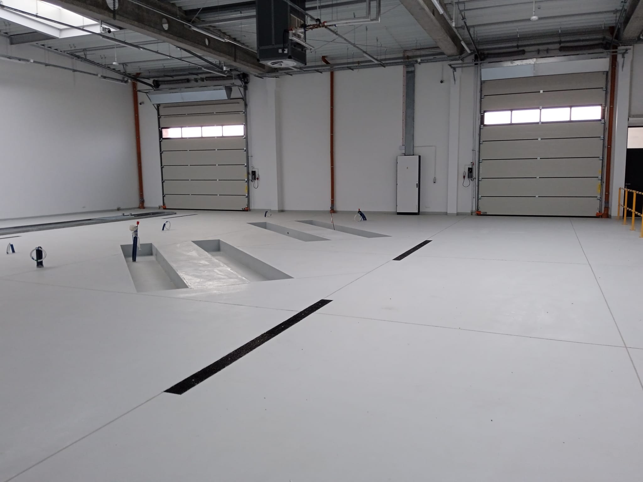 Jasne, przestronne wnętrze hali z dwoma bramami segmentowymi i kanałami rewizyjnymi w posadzce. Widoczne instalacje pod sufitem i pionowe rury przy ścianach.
