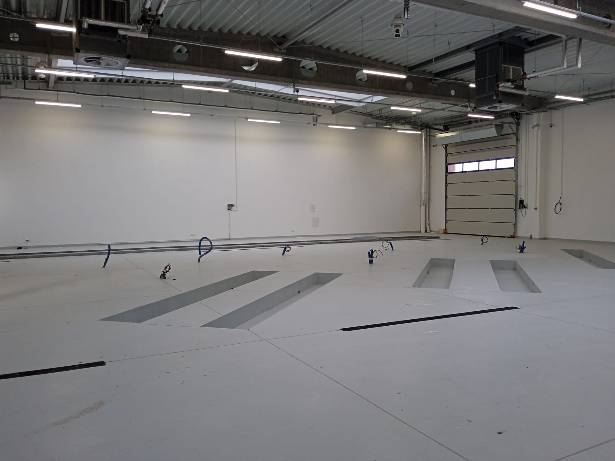 Wnętrze hali warsztatowej z kanałami rewizyjnymi w posadzce, oświetleniem sufitowym i bramą segmentową. Widoczne instalacje i elementy wyposażenia.