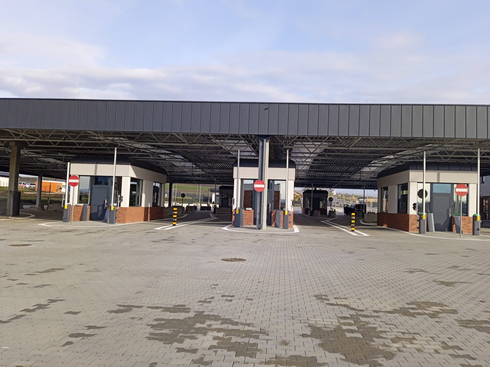 Nowoczesny punkt kontroli granicznej z szarym zadaszeniem i kostką brukową. Widoczne budki kontrolne z ceglanymi elementami i znakami zakazu wjazdu. Jasne niebo w tle.