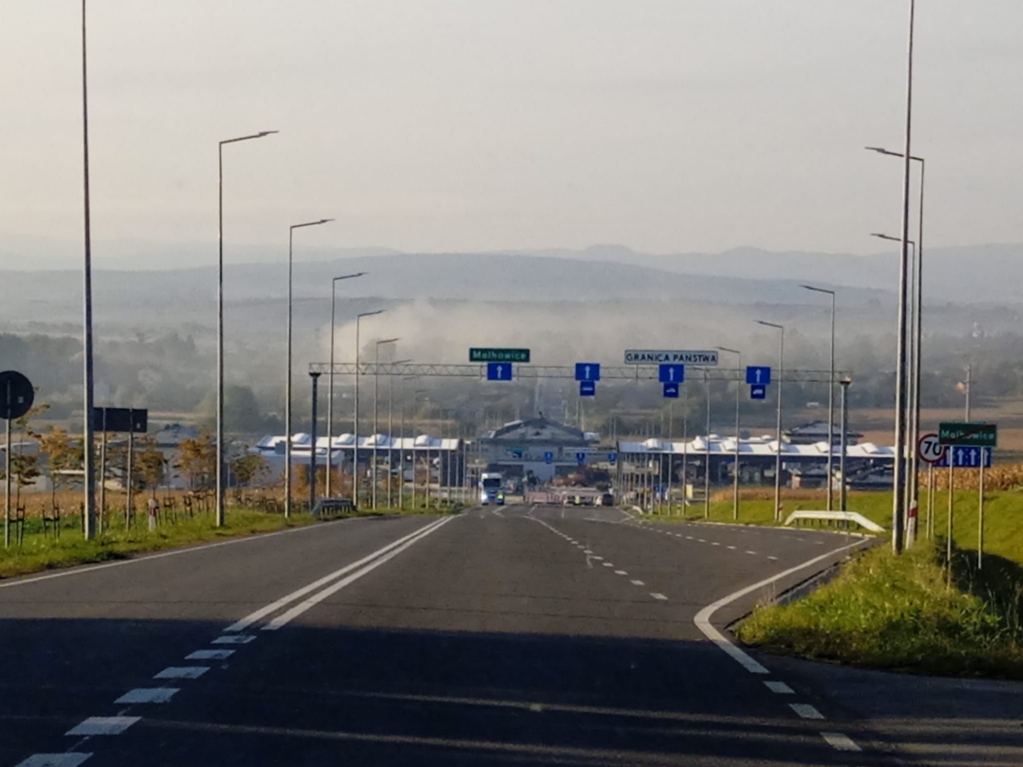 Asfaltowa droga prowadząca do przejścia granicznego z budynkami kontrolnymi i szlabanami, otoczona polami i górami w oddali, oświetlona porannym światłem.