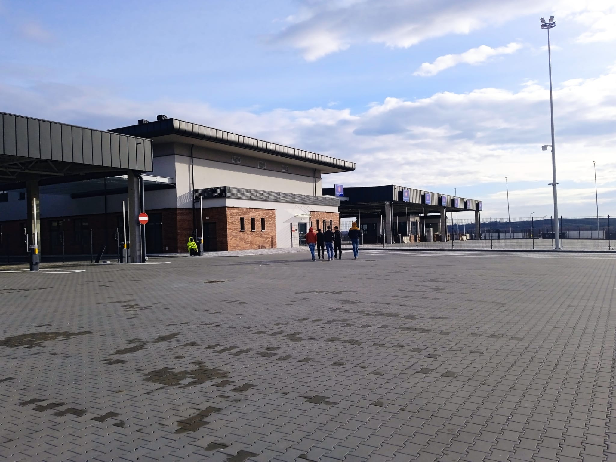 Rozległy plac wyłożony szarą kostką brukową, z budynkami w tle i grupą osób przechodzących przez przejście dla pieszych. Widoczne słupy oświetleniowe i błękitne niebo.