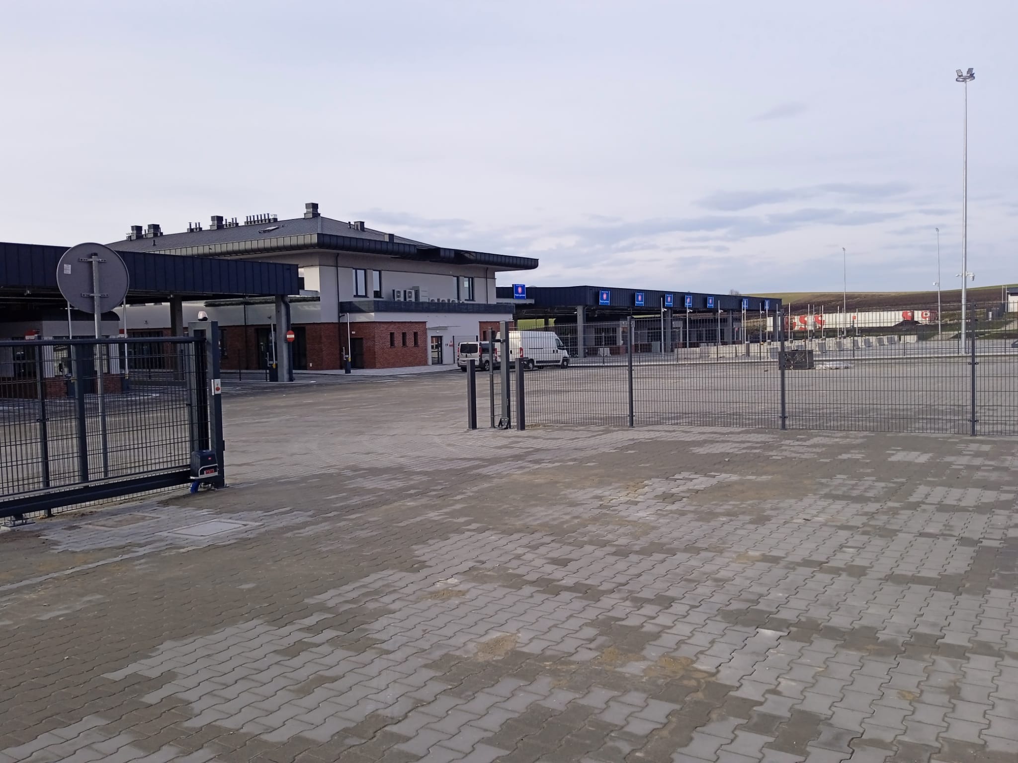 Rozległy plac wyłożony szarą kostką brukową, z budynkiem w tle. Widoczne bramy wjazdowe i szlabany. Całość w stonowanej kolorystyce, przy pochmurnym niebie.