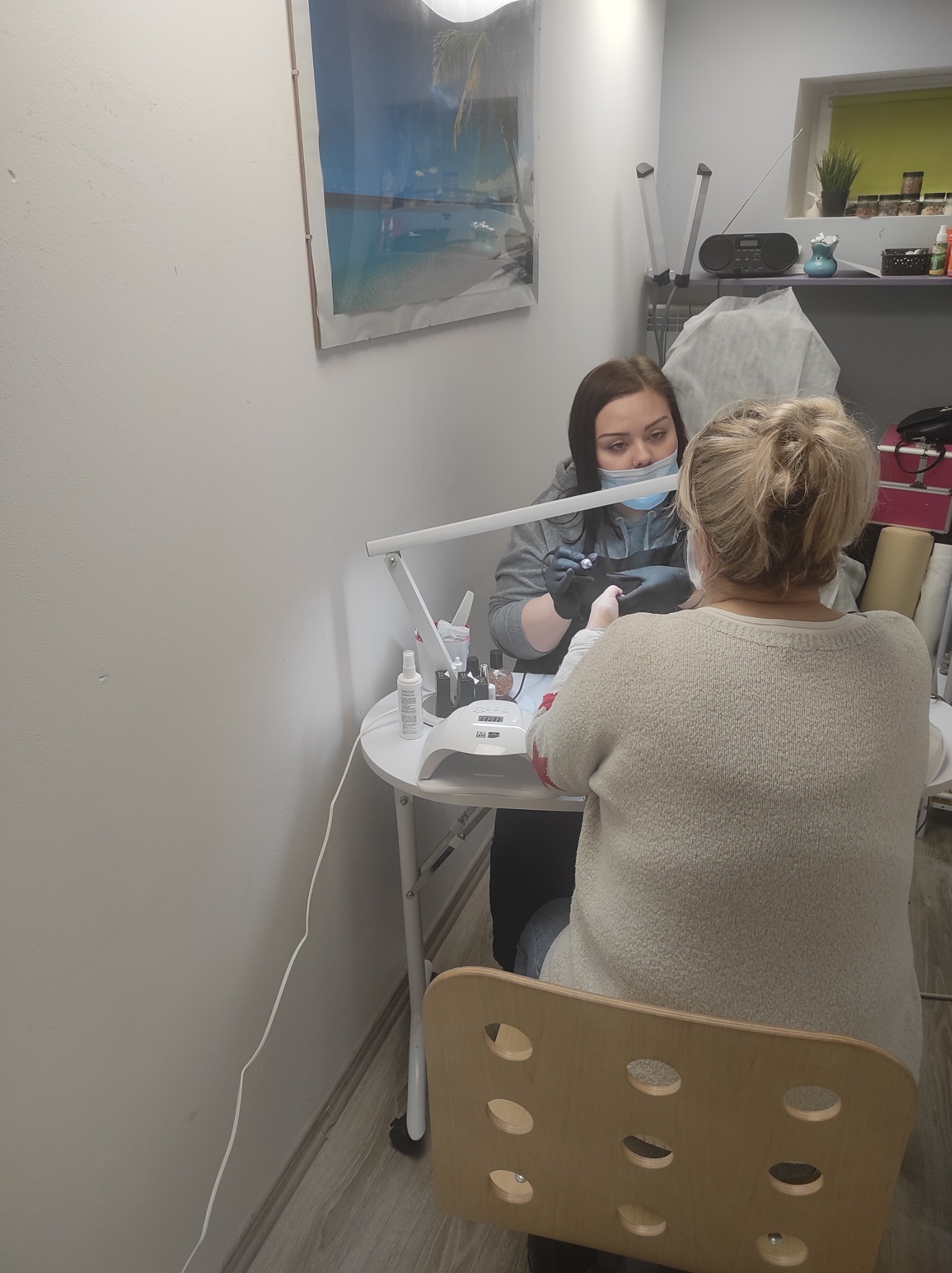 Stylistka paznokci w czarnych rękawiczkach i maseczce higienicznej precyzyjnie opracowuje paznokcie klientki siedzącej przy białym stole pod lampą, w tle widoczna półka z kosmetykami i obrazek...