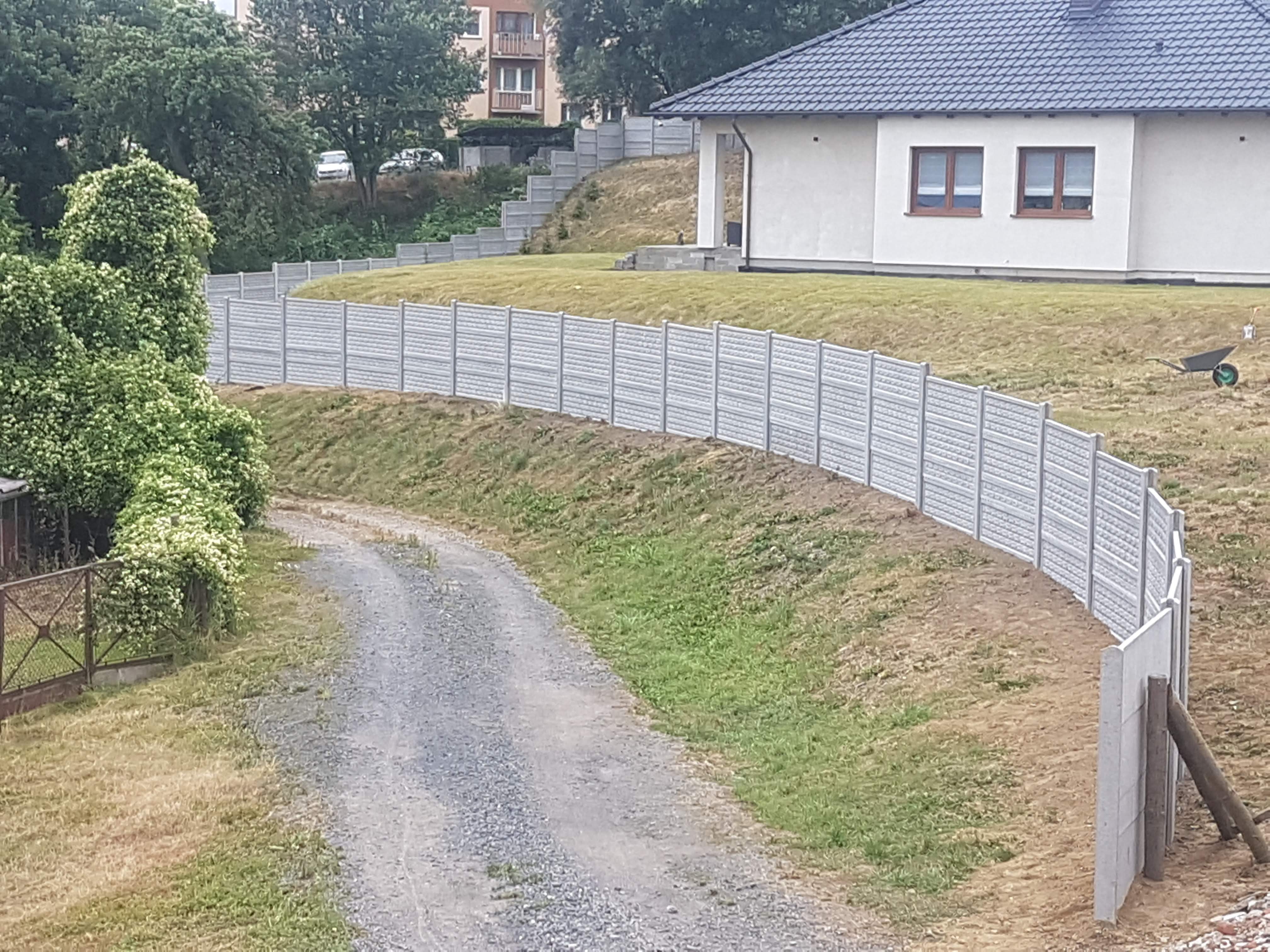 Długa, szara, betonowa konstrukcja oddzielająca posesję od drogi gruntowej, w tle dom z szarym dachem i budynek mieszkalny.