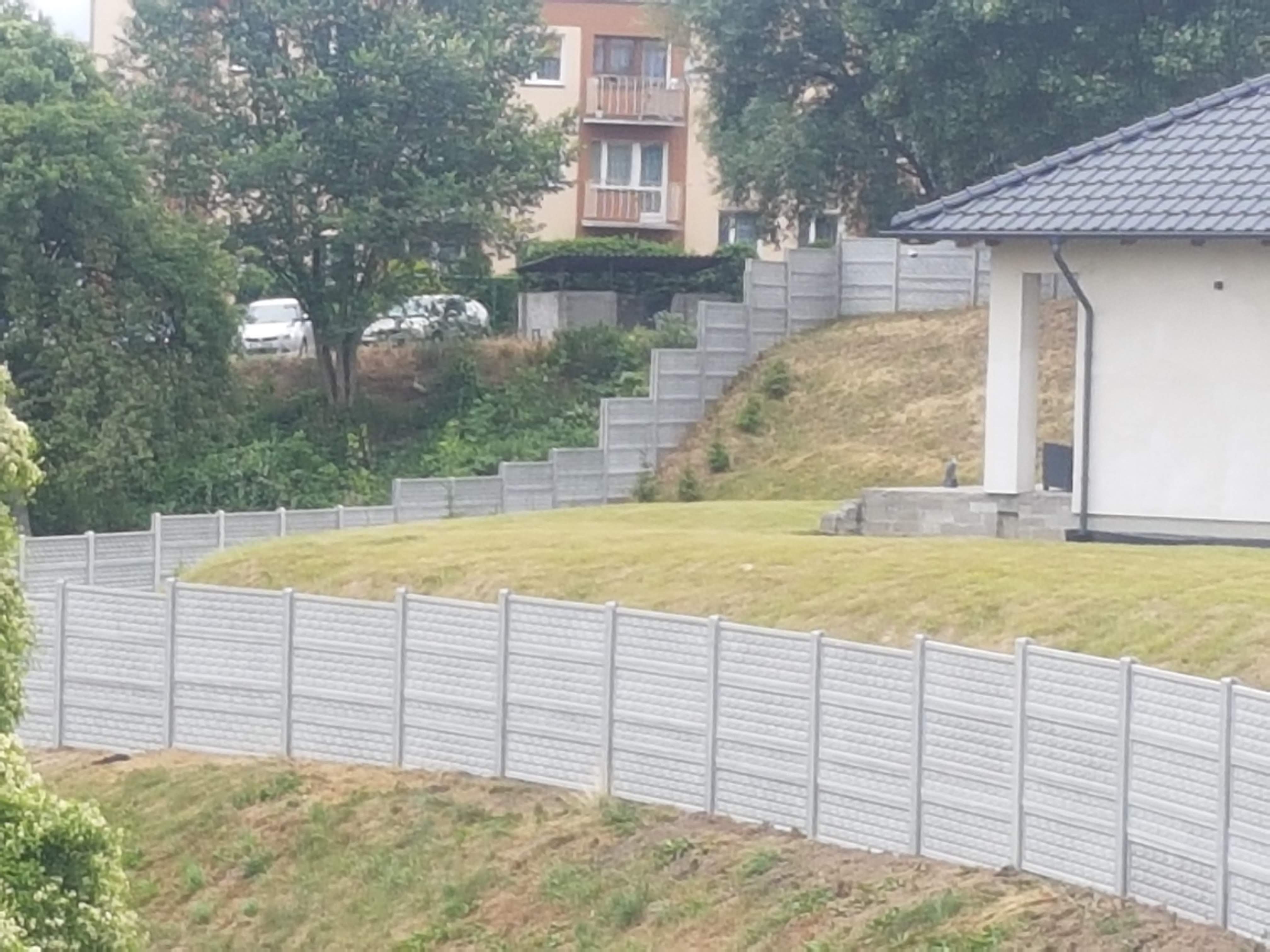 Długi, szary mur z prefabrykowanych elementów betonowych, wzniesiony na nierównym terenie, z budynkiem mieszkalnym i zielenią w tle.