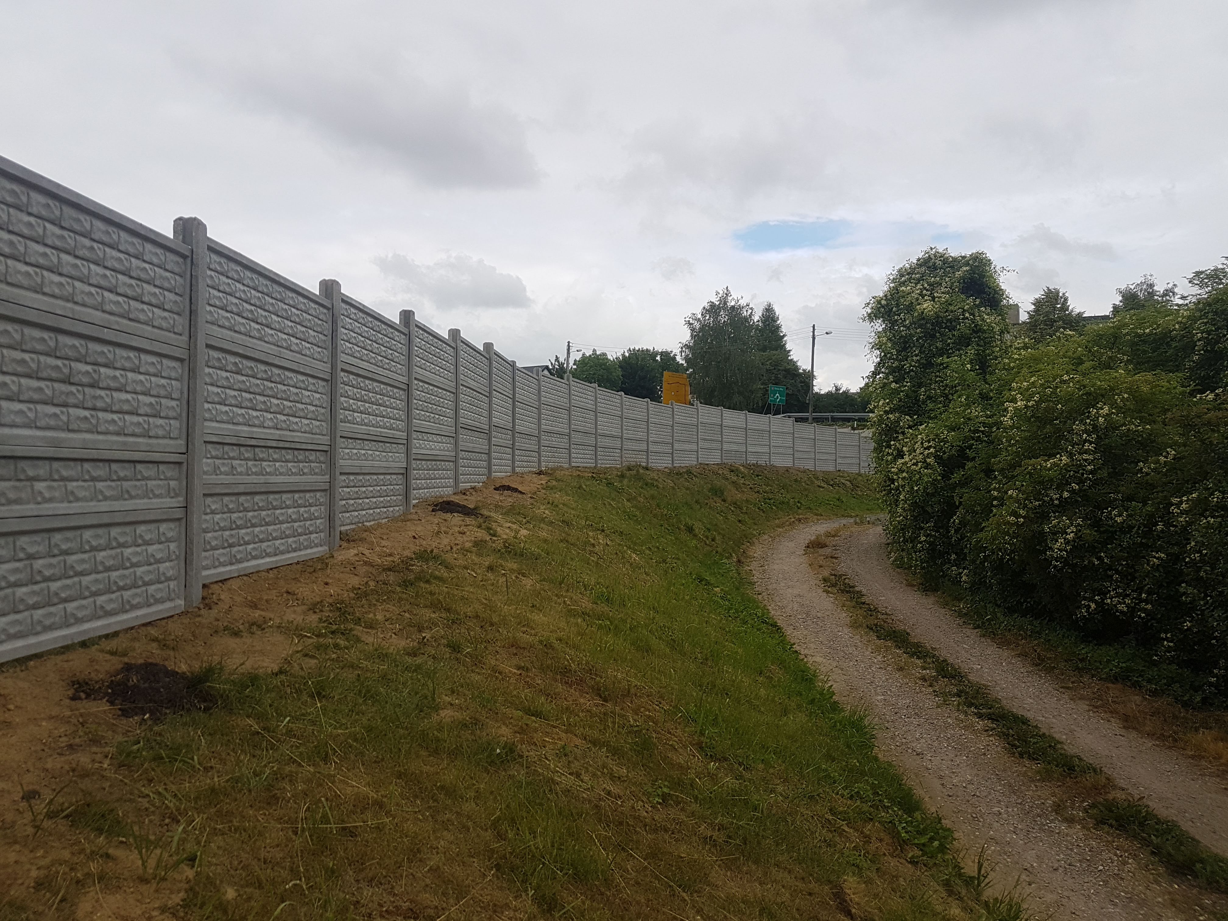 Długa, szara zapora akustyczna z prefabrykatów betonowych, ciągnąca się wzdłuż polnej drogi, za którą widać drogowskazy i zieleń drzew.