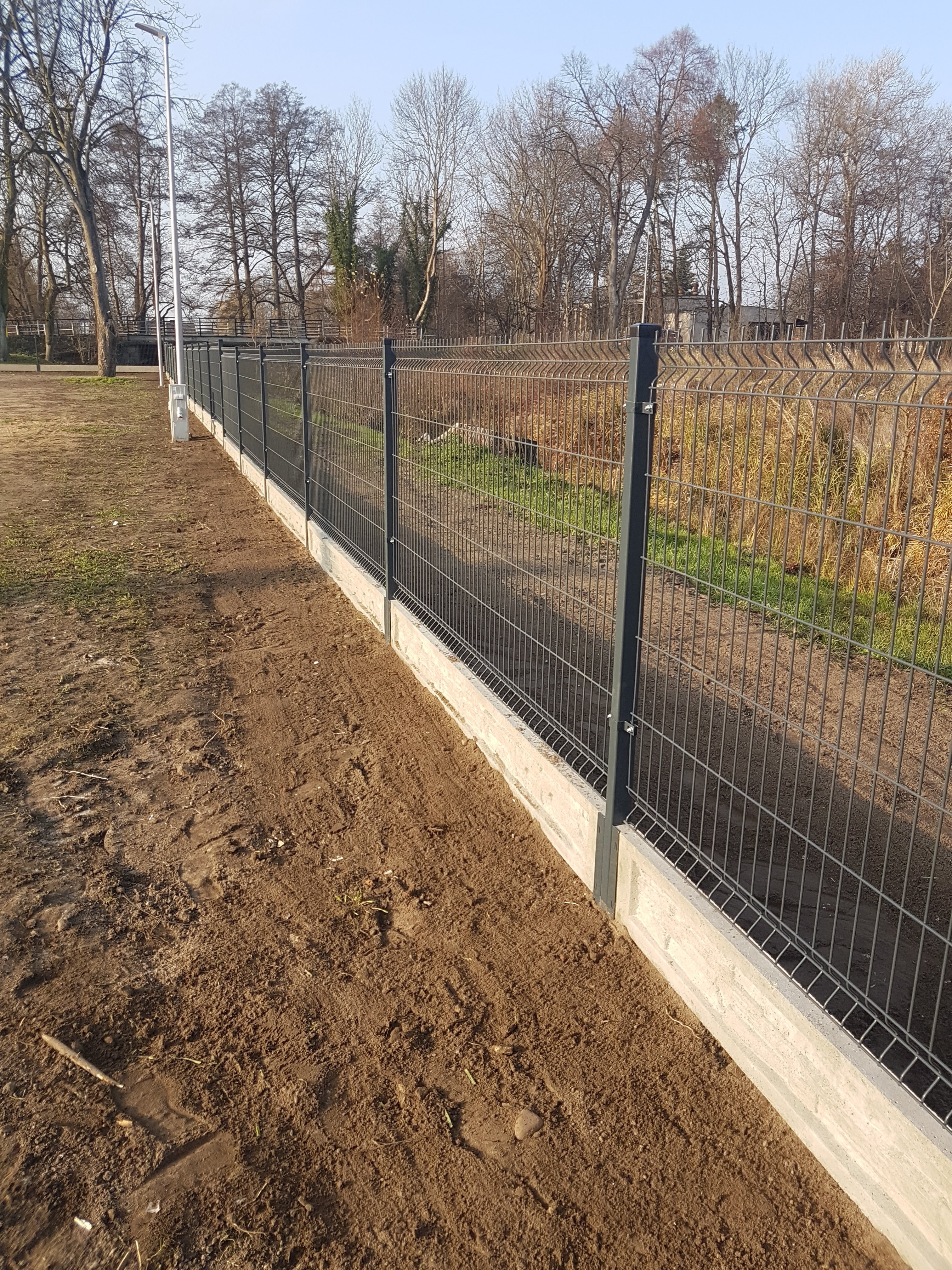 Nowo postawione, szare ogrodzenie panelowe z podmurówką betonową na niezagospodarowanym terenie z widocznymi śladami prac ziemnych.