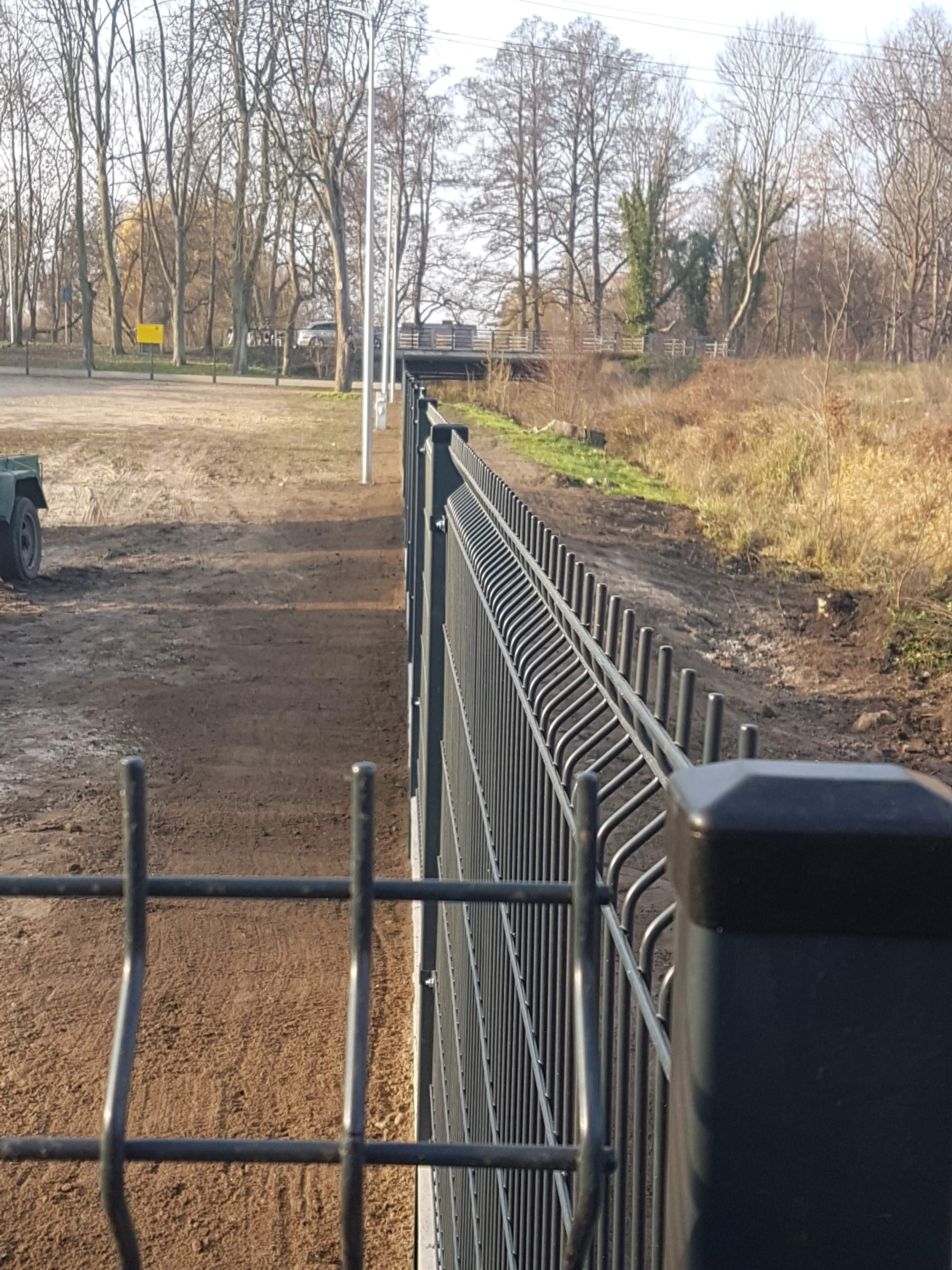 Nowo postawione, ciemne ogrodzenie panelowe na wyrównanym terenie z widocznymi śladami opon, w tle drzewa i most.