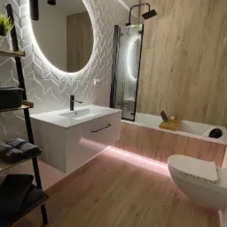 Nowoczesna łazienka z podświetlanym lustrem, białą umywalką na szafce, drewnopodobnymi płytkami na ścianach i podłodze oraz dekoracyjną drabinką na ręczniki.