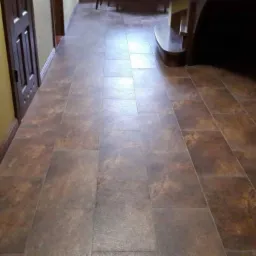 Ułożona brązowa podłoga z płytek imitujących kamień w korytarzu z ciemnymi drzwiami i fragmentem ceglanej ściany.