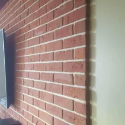 Dekoracyjna ściana z czerwonej cegły z białymi fugami, oświetlona punktowymi lampami sufitowymi, sąsiadująca z gładką, jasną ścianą.