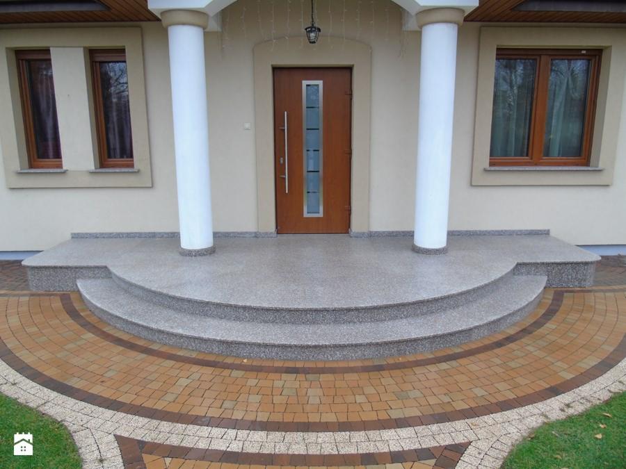 Wejście do domu z granitowymi schodami i półokrągłym chodnikiem z kostki brukowej w odcieniach beżu i brązu, otoczone trawnikiem.