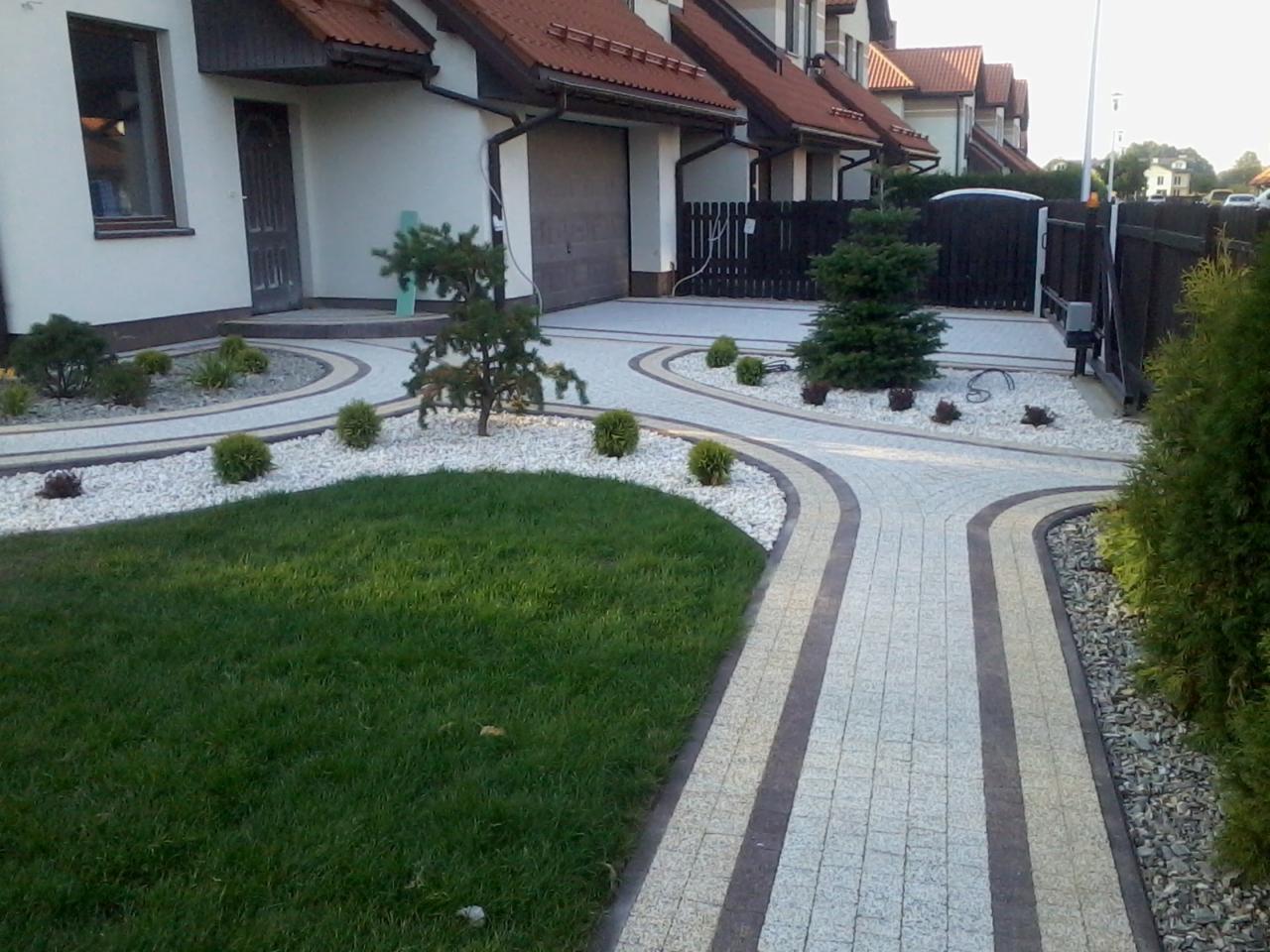 Ułożona kostka granitowa tworząca ścieżki i podjazd przed domem z białymi kamykami i zielonym trawnikiem, widoczne niskie krzewy i małe drzewka. W tle segment zabudowy szeregowej.