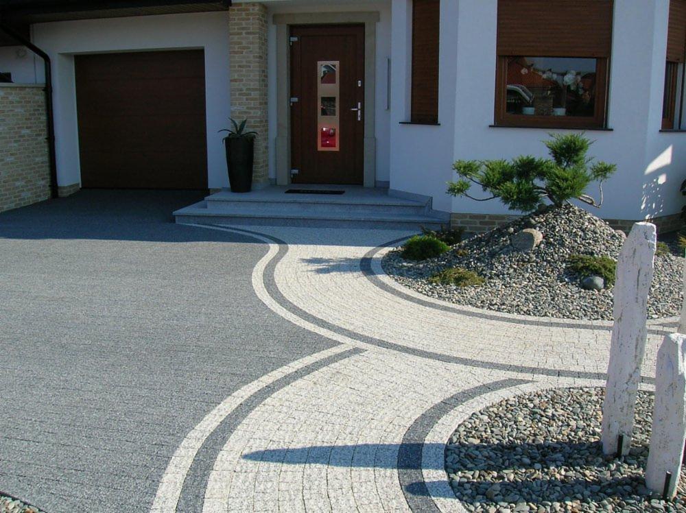 Ułożona kostka granitowa w formie wijącej się ścieżki z szarego i jasnego granitu, prowadząca do domu z brązowymi drzwiami i garażem, obok skalniak z małą sosną i kamieniami.