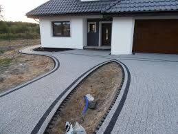 Ułożona kostka brukowa w odcieniach szarości z czarnymi obrzeżami tworząca ścieżkę i ozdobny element przed nowoczesnym domem z białymi ścianami i brązową bramą garażową. Fragment rabaty z ziemią...