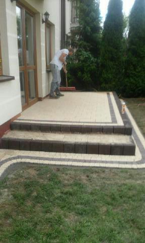 Pracownik zamiata nowo ułożoną kostkę na tarasie z dwoma stopniami, wykończoną ciemniejszym obramowaniem, przy domu z dużymi oknami i zielonym trawnikiem.