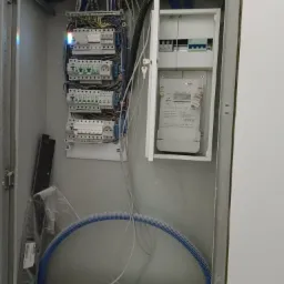 Otwarta skrzynka elektryczna z zamontowanymi bezpiecznikami, licznikiem energii i przewodami, niebieska rura karbowana w dolnej części.