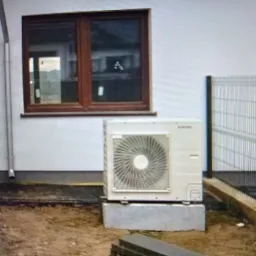 Zewnętrzna jednostka klimatyzacji zamontowana na betonowych bloczkach przy ścianie budynku z brązowymi oknami i drzwiami, widoczny fragment ogrodzenia.