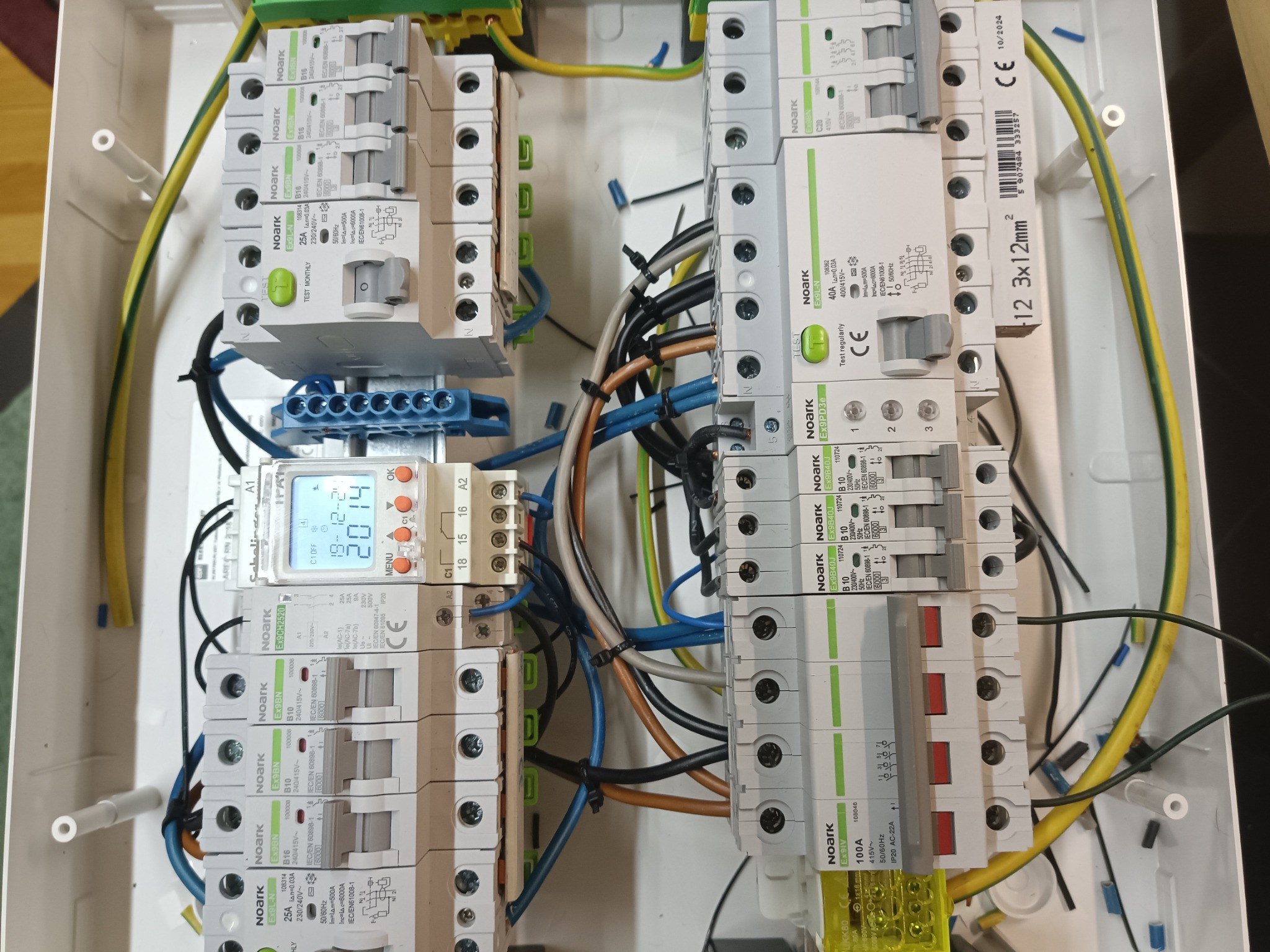 Rozdzielnica elektryczna z zamontowanymi bezpiecznikami i wyłącznikami różnicowoprądowymi marki Noark, widoczne przewody w różnych kolorach oraz programator czasowy z wyświetlaną datą 19-12-2014.