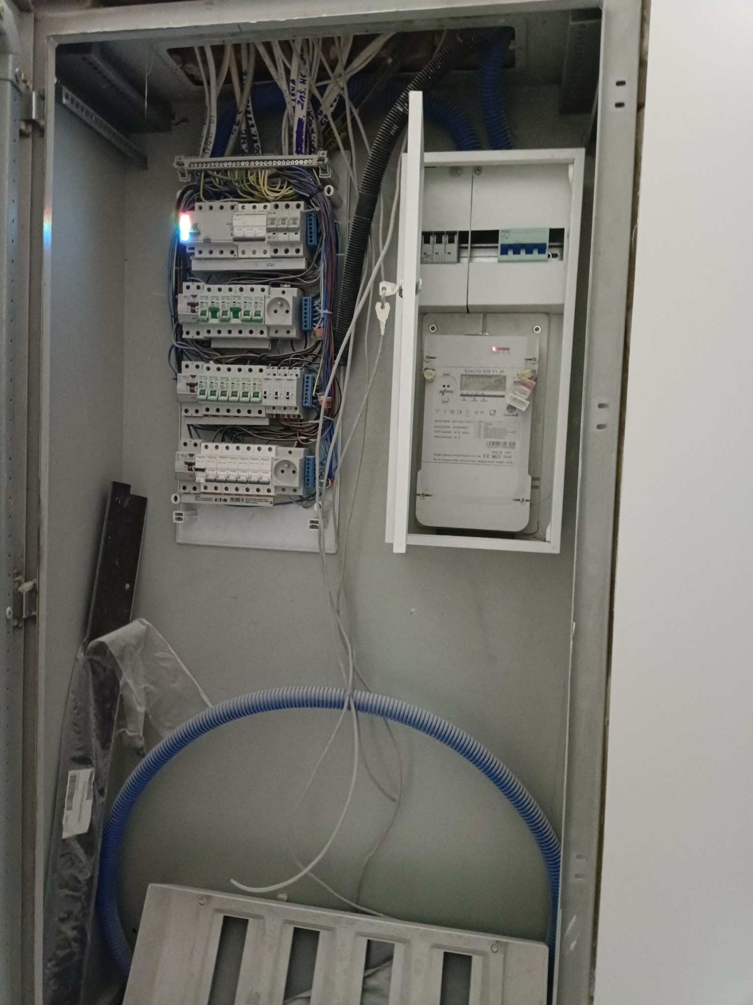 Otwarta skrzynka elektryczna z zamontowanymi bezpiecznikami, licznikiem energii i przewodami, niebieska rura karbowana w dolnej części.