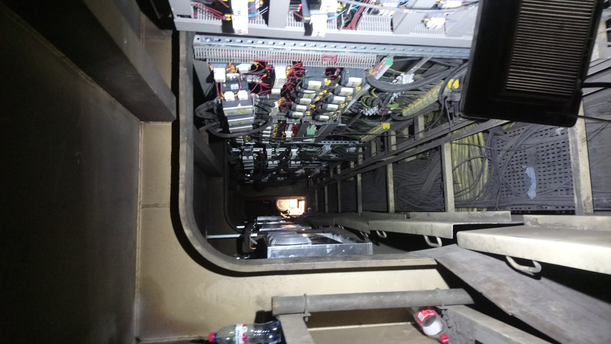 Wnętrze szybu z widocznymi kablami, rozdzielnicami i metalowymi konstrukcjami; butelka wody i jeden but na podłodze.