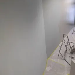 Świeżo zagruntowana ściana z widocznymi śladami szpachlowania, zabezpieczone schody i podłoga folią malarską, drabina malarska w rogu pomieszczenia.