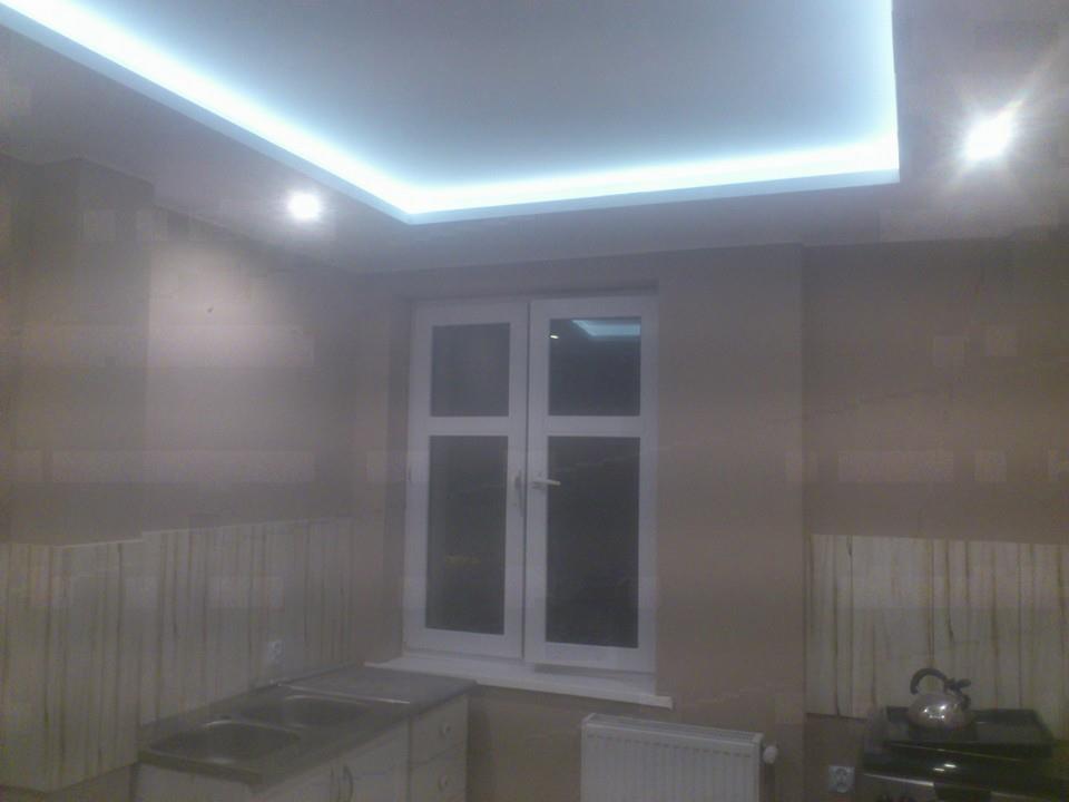 Wykończona łazienka z beżowymi płytkami, białym oknem i podwieszanym sufitem z oświetleniem LED, widoczny fragment szafki pod umywalkę i grzejnik.