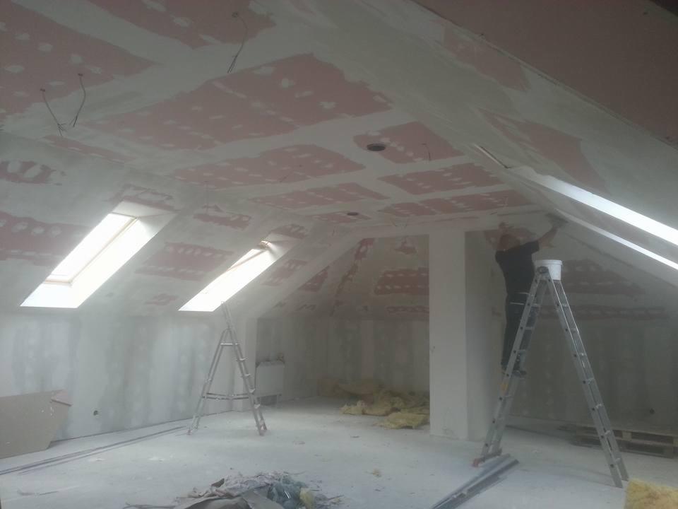 Poddasze w trakcie prac wykończeniowych: mężczyzna na drabinie szpachluje skośny sufit przy oknie dachowym, widoczne ślady po łatach gipsowych na suficie, w tle druga drabina i materiały budowlane.