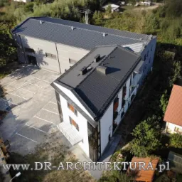 Widok z lotu ptaka na nowoczesny budynek mieszkalny z czarnym dachem i białymi ścianami, przylegający do budynku usługowego z parkingiem, otoczony zielenią.