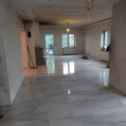 MICHAŁ K.MMBUD Michał Kirstajn - Wnętrze w trakcie prac wykończeniowych: świeżo położone, błyszczące, szare płytki podłogowe imitujące marmur, surowe ściany wymagające wykończenia, widoczne kable zwisające z sufitu, okna...