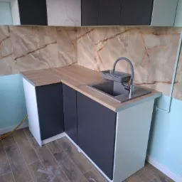 Nowoczesna zabudowa kuchenna z blatem imitującym drewno, szarymi szafkami i marmurową mozaiką na ścianie. Uwagę zwraca nietypowe połączenie kolorów i materiałów.