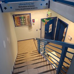 MICHAŁ K.MMBUD Michał Kirstajn - Klatka schodowa w trakcie odświeżania: świeżo pomalowane ściany w jasnym odcieniu, zabezpieczone taśmą schody i framugi drzwi, błękitne barierki i elementy dekoracyjne.