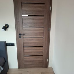 MICHAŁ K.MMBUD Michał Kirstajn - Zamknięte, nowoczesne drzwi wewnętrzne z ciemnego drewna z poziomymi wstawkami, czarną klamką i widocznymi zawiasami, zamontowane w jasnym pomieszczeniu.