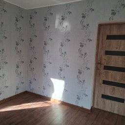 MICHAŁ K.MMBUD Michał Kirstajn - Pokój po remoncie: ściany w jasnej tapecie z motywem roślinnym, drewniana podłoga i drzwi z przeszkleniami. Widoczne listwy przypodłogowe.