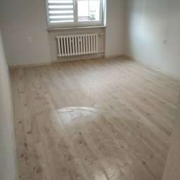 MICHAŁ K.MMBUD - Jasne panele podłogowe ułożone w pustym pokoju z białymi ścianami, widoczne okno z żaluzjami i grzejnik.