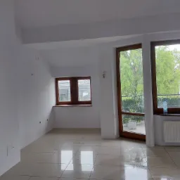 Jasne, przestronne wnętrze po malowaniu ścian na biało, z widocznymi dwoma oknami z drewnianą ramą, beżową podłogą z płytek i białym grzejnikiem pod oknem balkonowym.