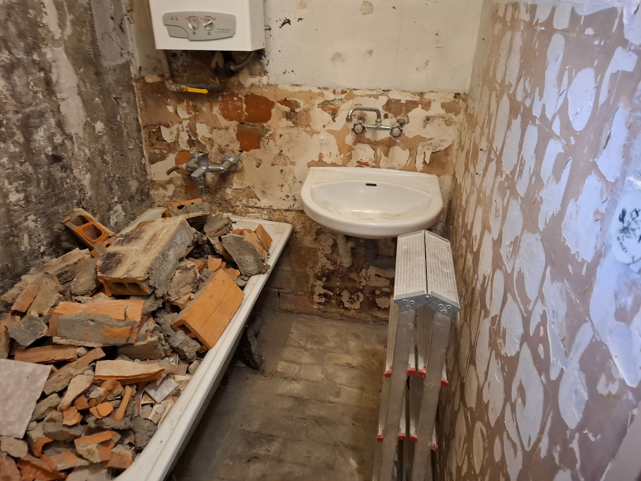 Łazienka w trakcie demontażu: gruz ceglany w wannie, odsłonięte fragmenty muru, stara umywalka i drabina aluminiowa przy ścianie z resztkami farby.