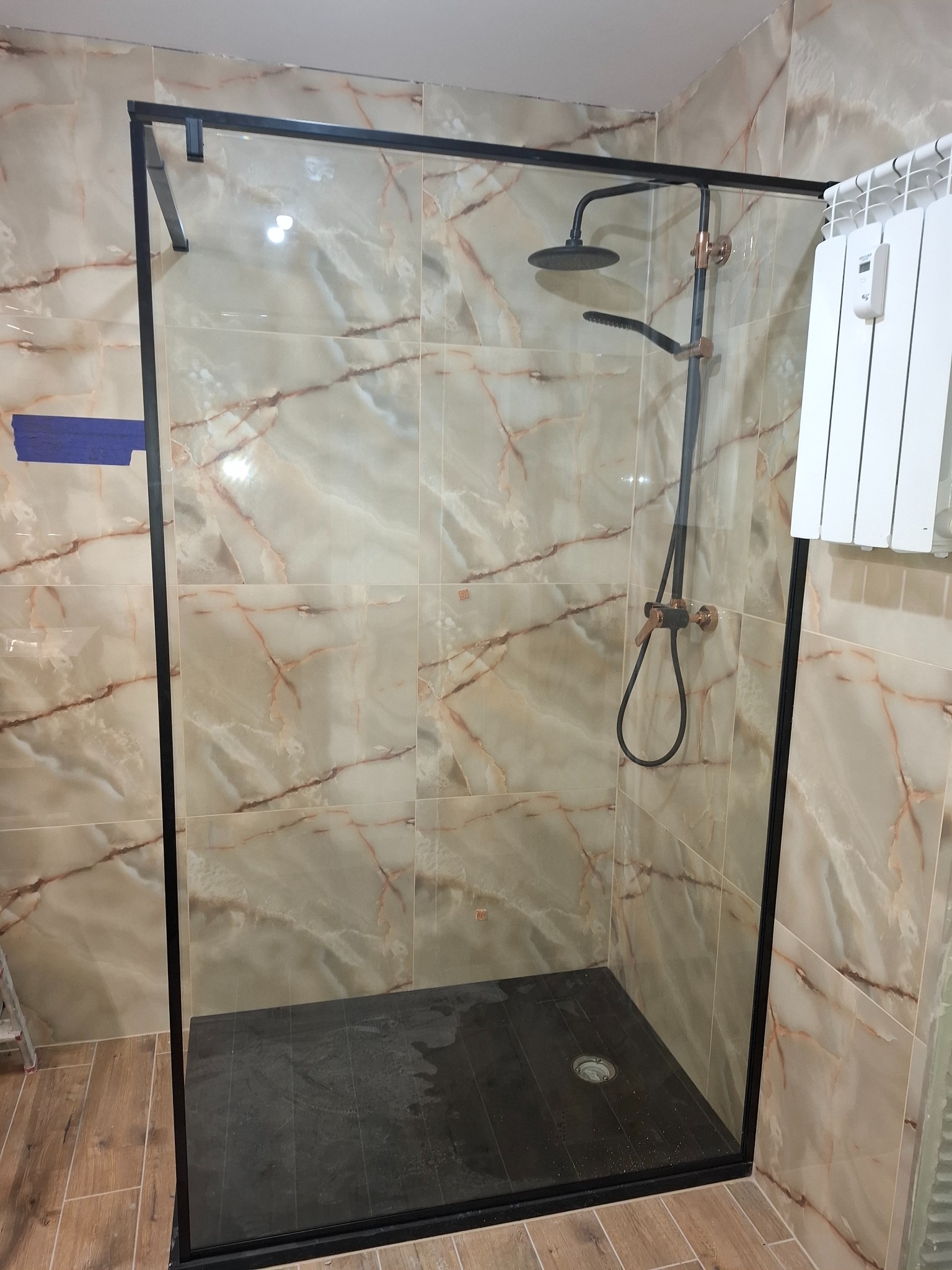 Nowoczesna kabina prysznicowa z czarną ramą i deszczownicą, wykończona marmurowymi płytkami. Widoczny grzejnik drabinkowy.