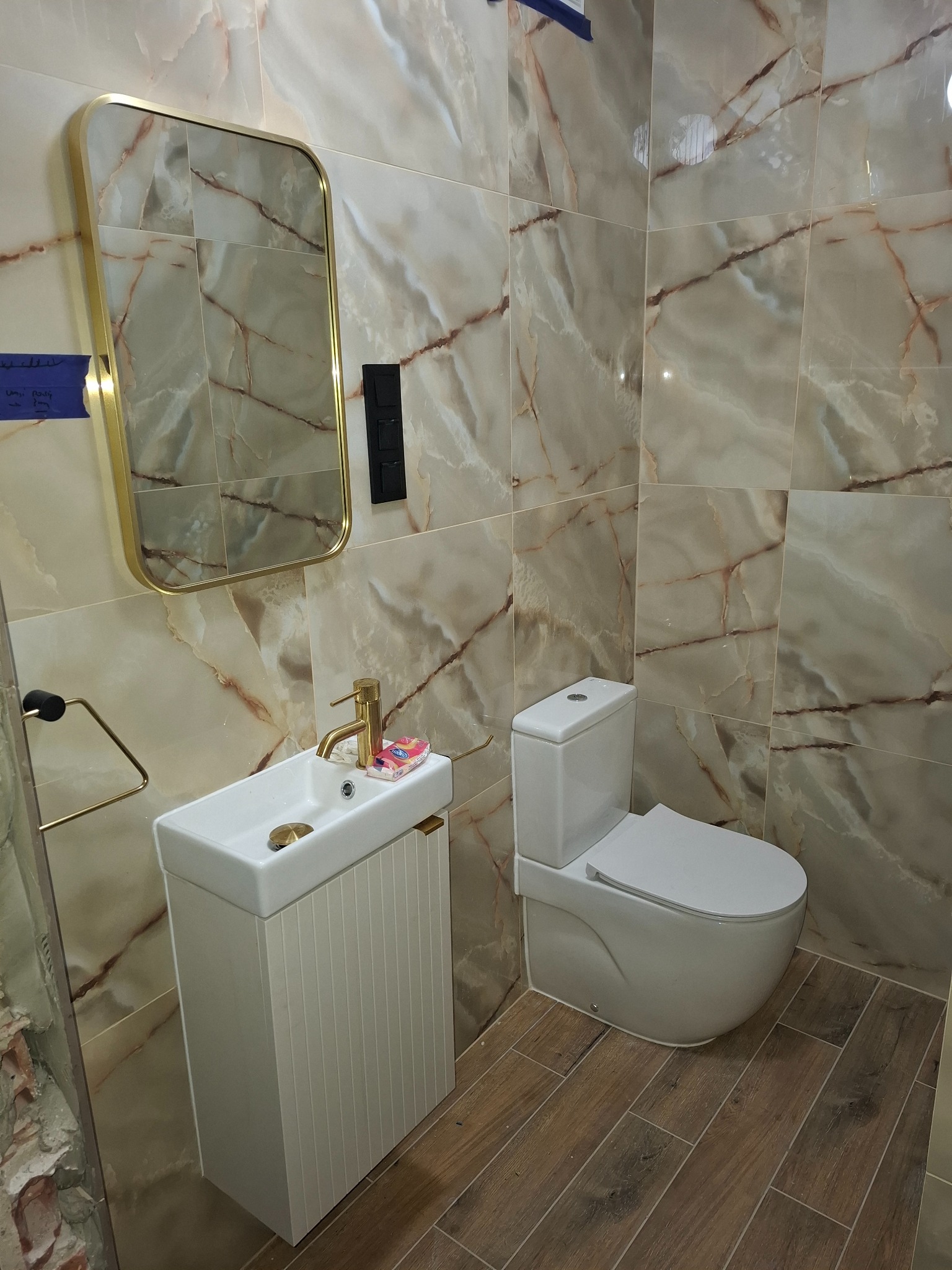 Wykończona łazienka z marmurowymi płytkami, złotą armaturą i drewnopodobną podłogą. Umywalka z szafką, lustro w złotej ramie, toaleta. Nowoczesny design.