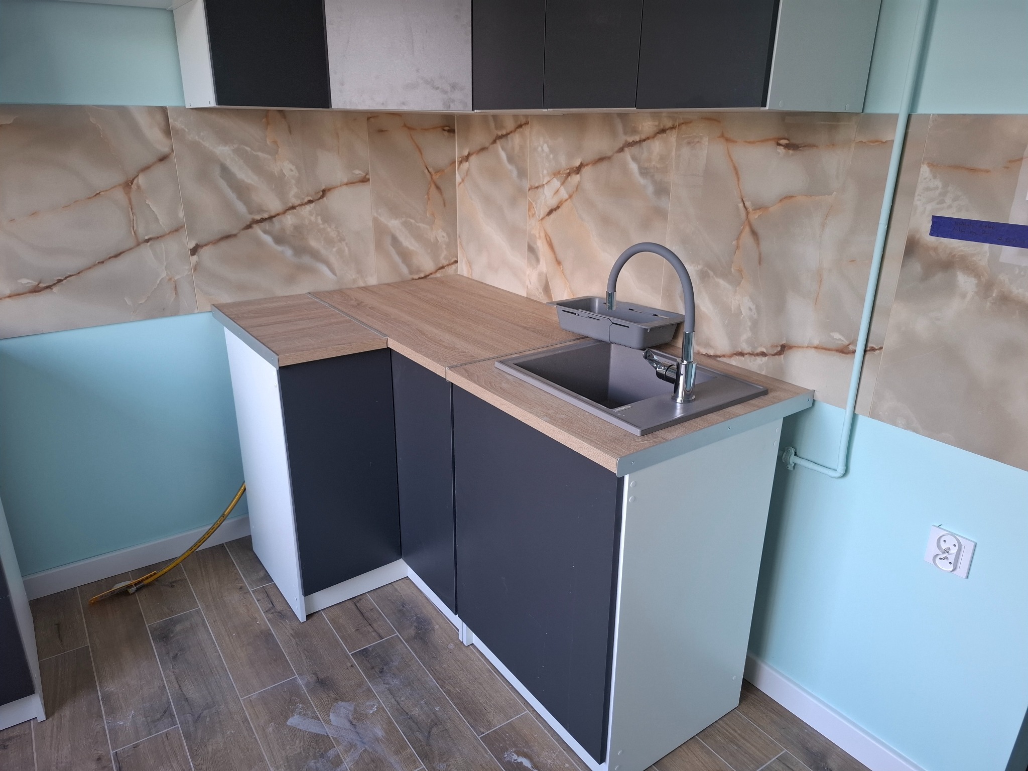 Nowoczesna zabudowa kuchenna z blatem imitującym drewno, szarymi szafkami i marmurową mozaiką na ścianie. Uwagę zwraca nietypowe połączenie kolorów i materiałów.