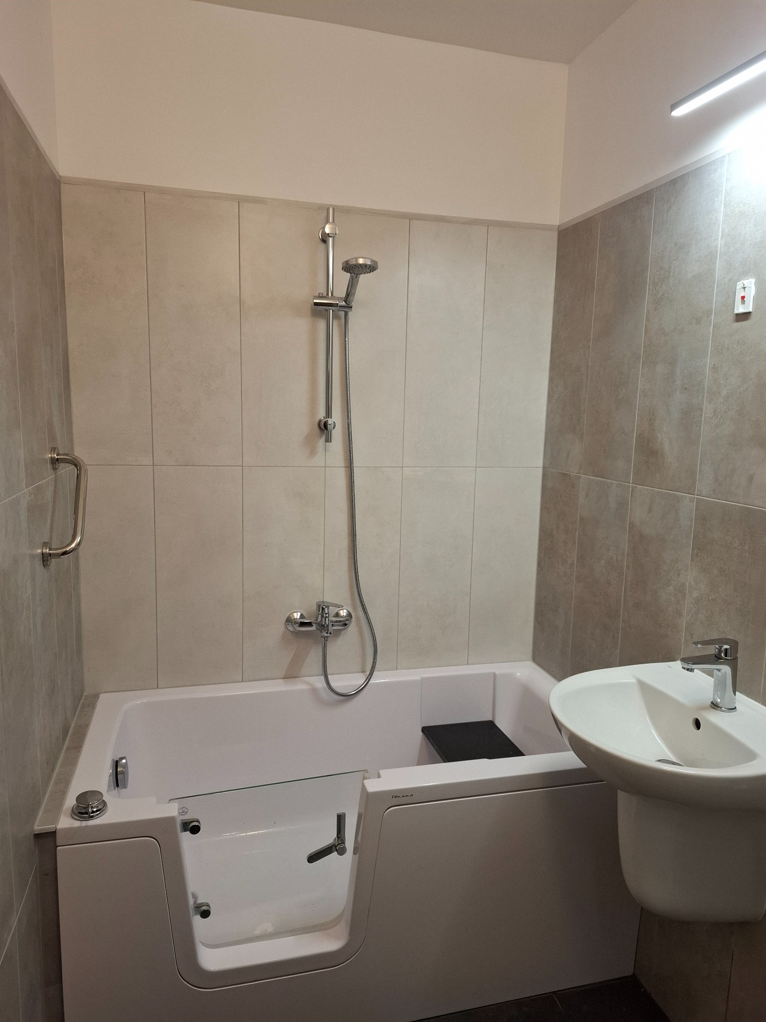 Wyremontowana łazienka z beżowymi płytkami, białą wanną z drzwiczkami i umywalką. Widoczny chromowany zestaw prysznicowy i uchwyt. Nowoczesne wykończenie.