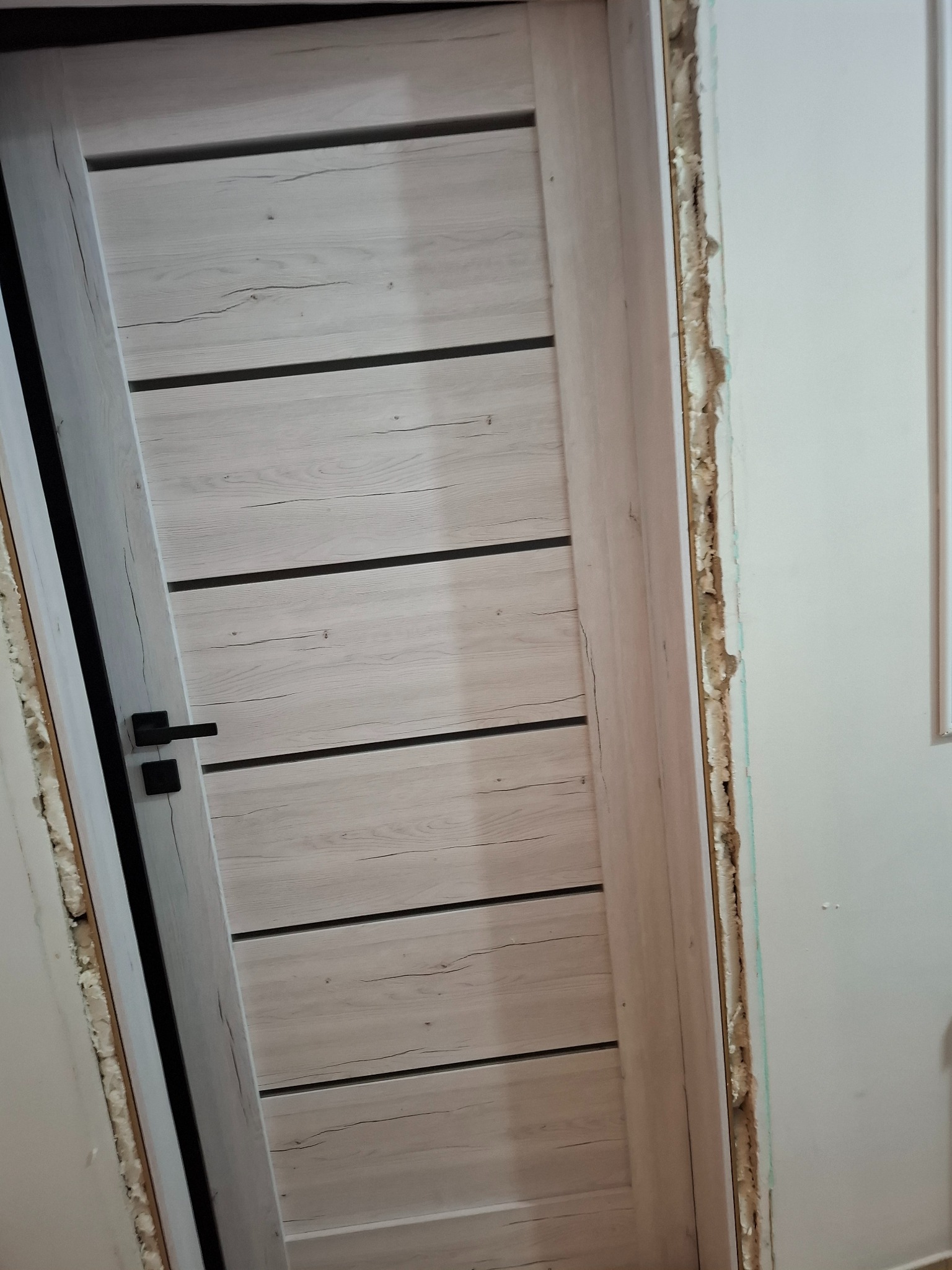 Montaż drzwi wewnętrznych: drewniane drzwi z czarną klamką, widoczne pianka montażowa i surowa ściana, co sugeruje etap prac wykończeniowych.