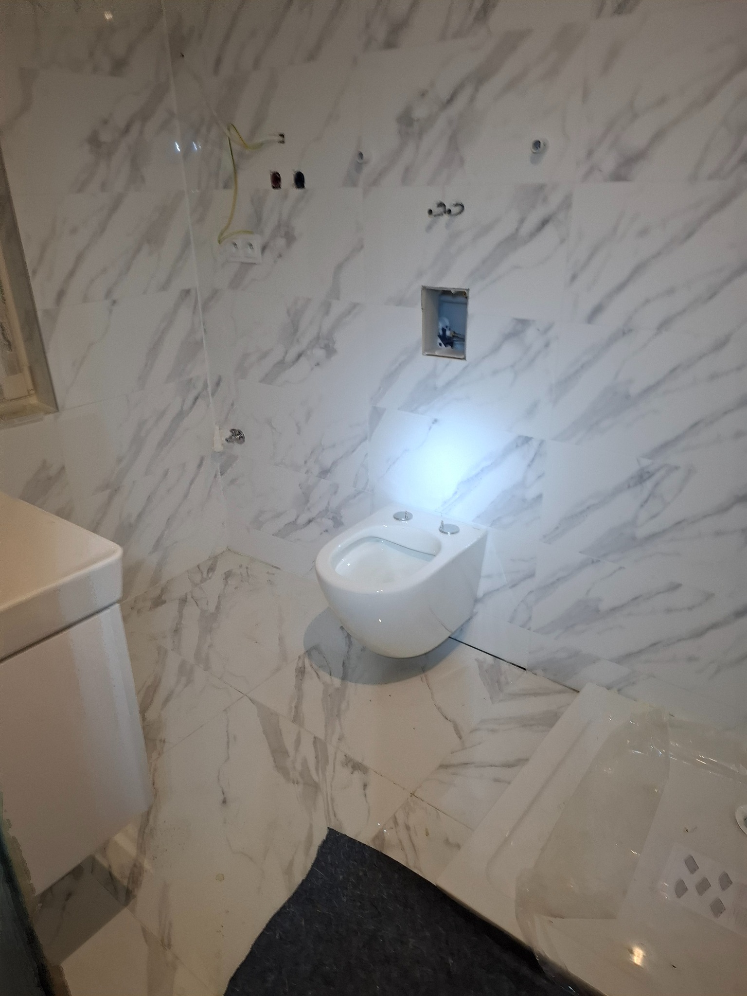 Biała, podwieszana toaleta w łazience wykończonej płytkami imitującymi marmur. Widoczne przygotowane instalacje i fragment wanny. Nowoczesny design.